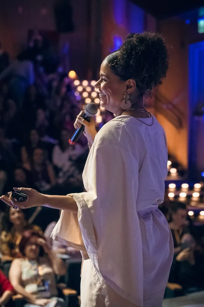Ediane Ribeiro de perfil no palco, falando ao microfone diante do público